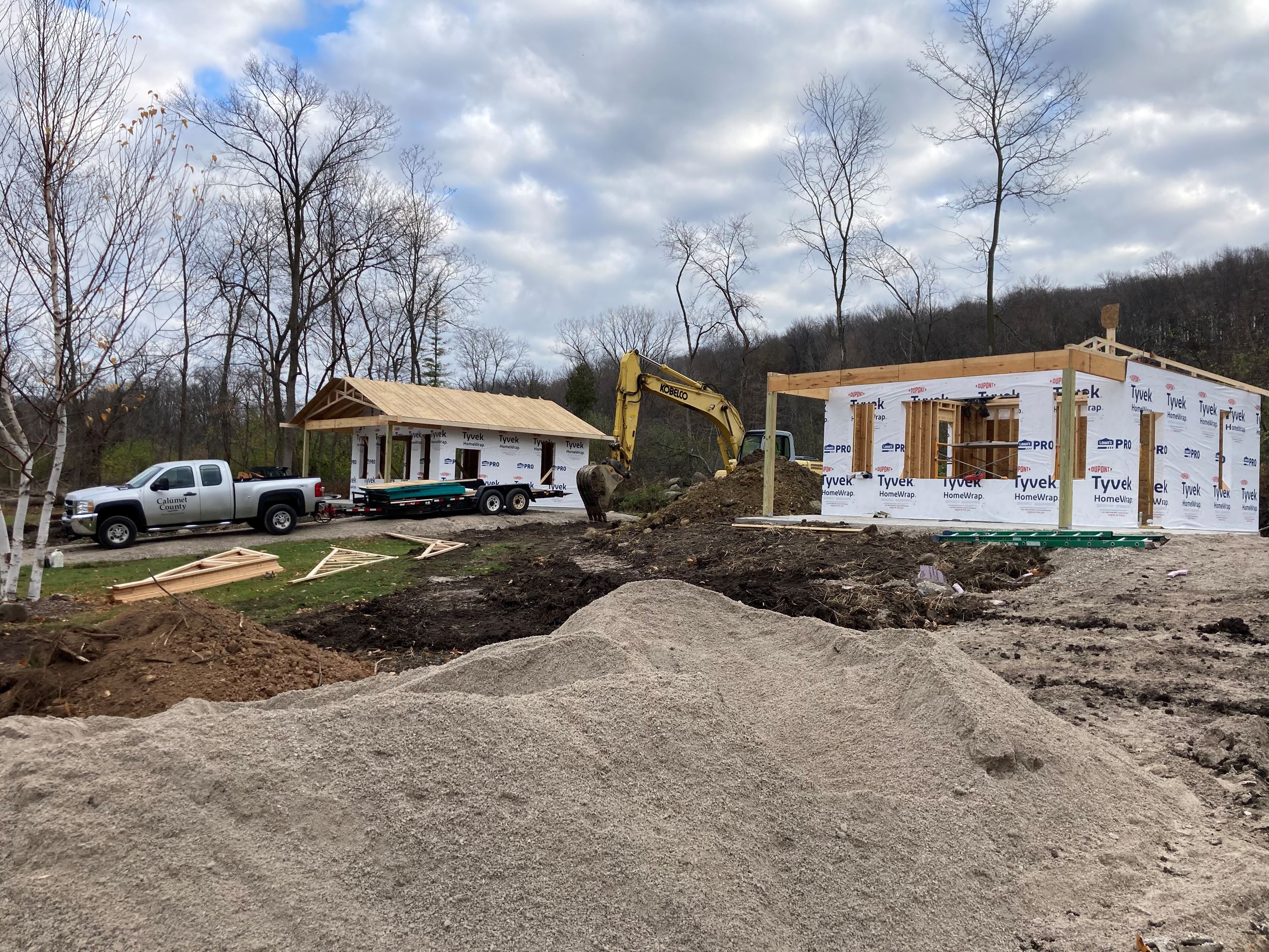 two partially constructed cabins with grey pickup truck and excavator in background
